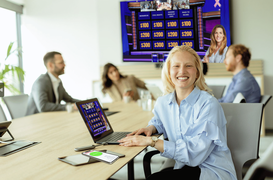 Woman sitting at a meeting table with 3 other people with a laptop that shows Jeoparty Social Women's Edition on the screen